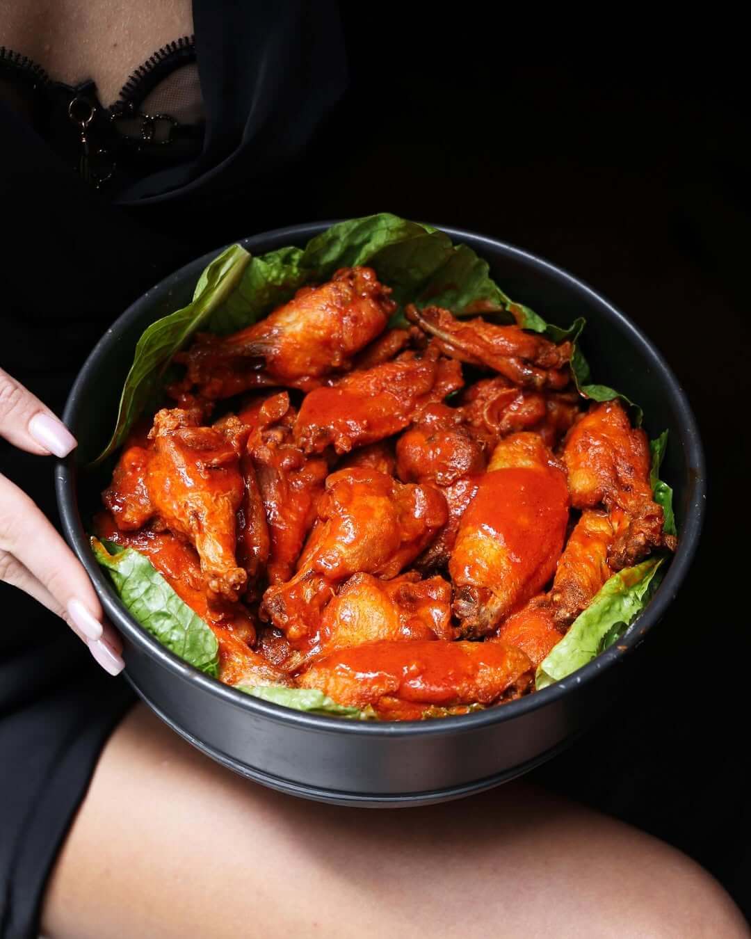 Bowl of spicy buffalo chicken wings on green lettuce leaves held by a person wearing black clothing.